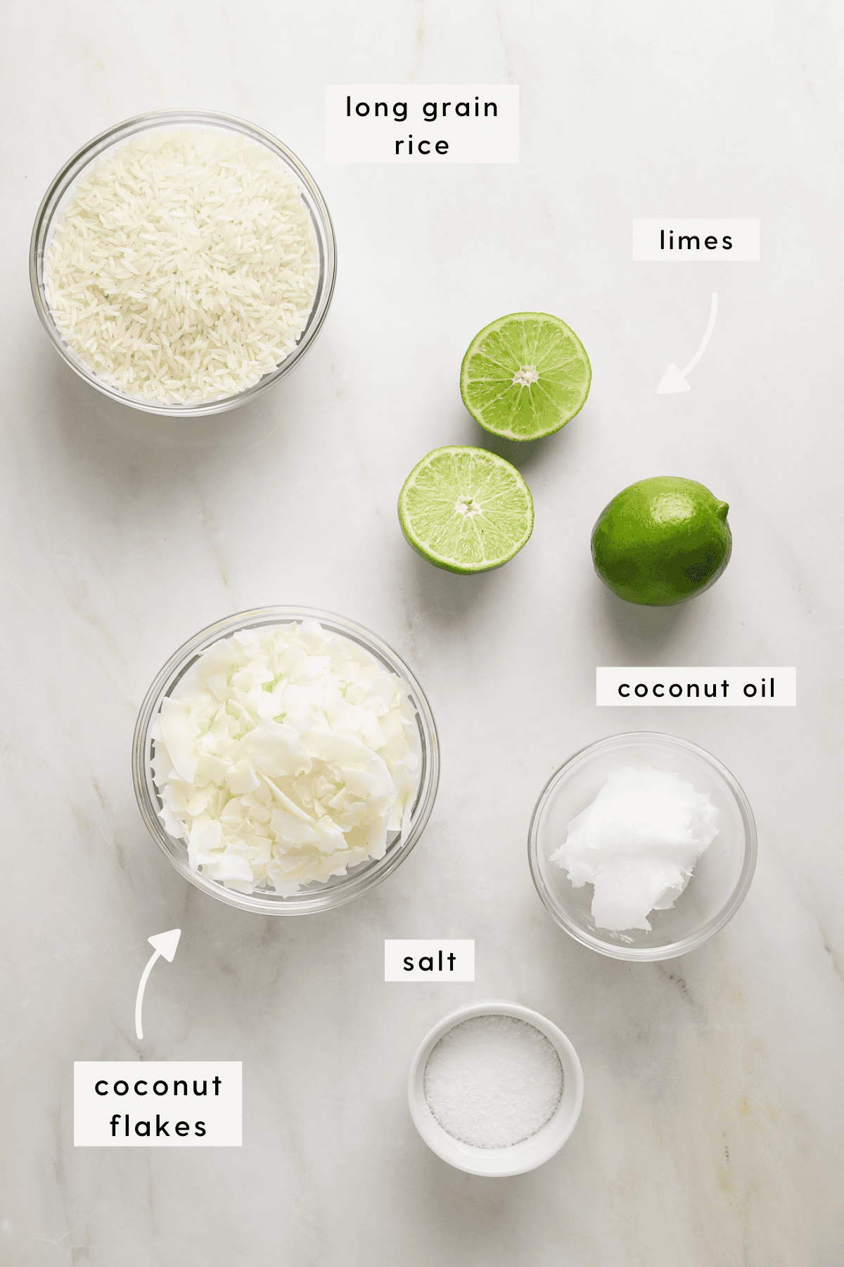 A small bowl of dried rice, three limes, coconut flakes and coconut oil on a marble tabletop.