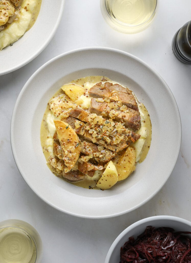 A bowl filled with a pork chop, apple and whole grain mustard sauce.