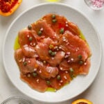 Thin slices of raw tuna on a white plate topped with capers, citrus zest and olive oil.