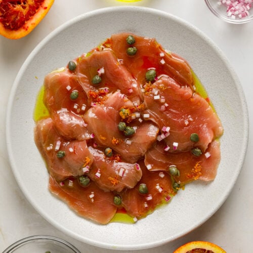 Thin slices of raw tuna on a white plate topped with capers, citrus zest and olive oil.