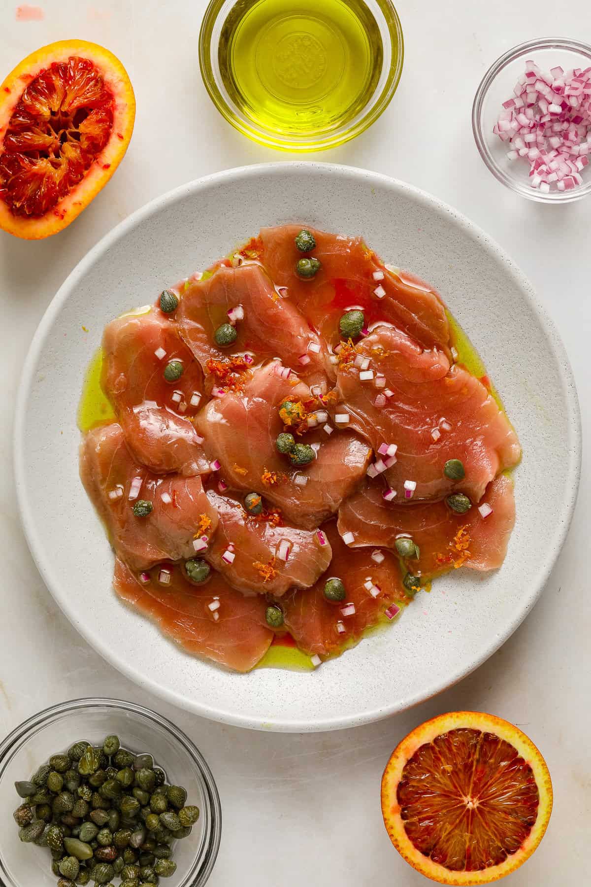 Thin slices of raw tuna on a white plate topped with capers, citrus zest and olive oil.