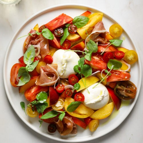 A large white platter filled with a stone fruit, tomato and burrata salad on a marble tabletop with two glasses of wine off to the side.