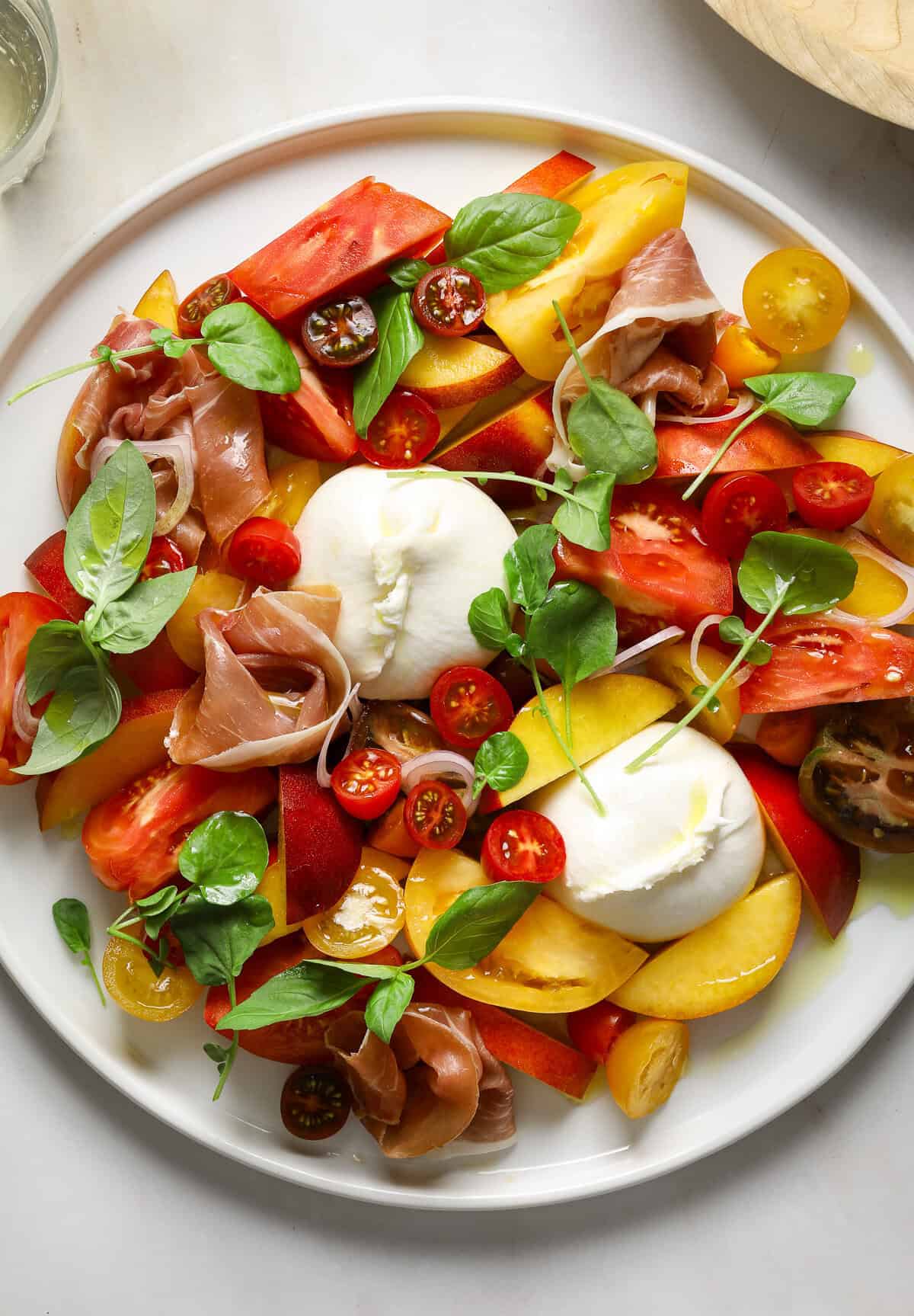 A large salad with slices of stone fruit, cherry tomatoes, fresh basil and mozzarella cheese on a white plate.