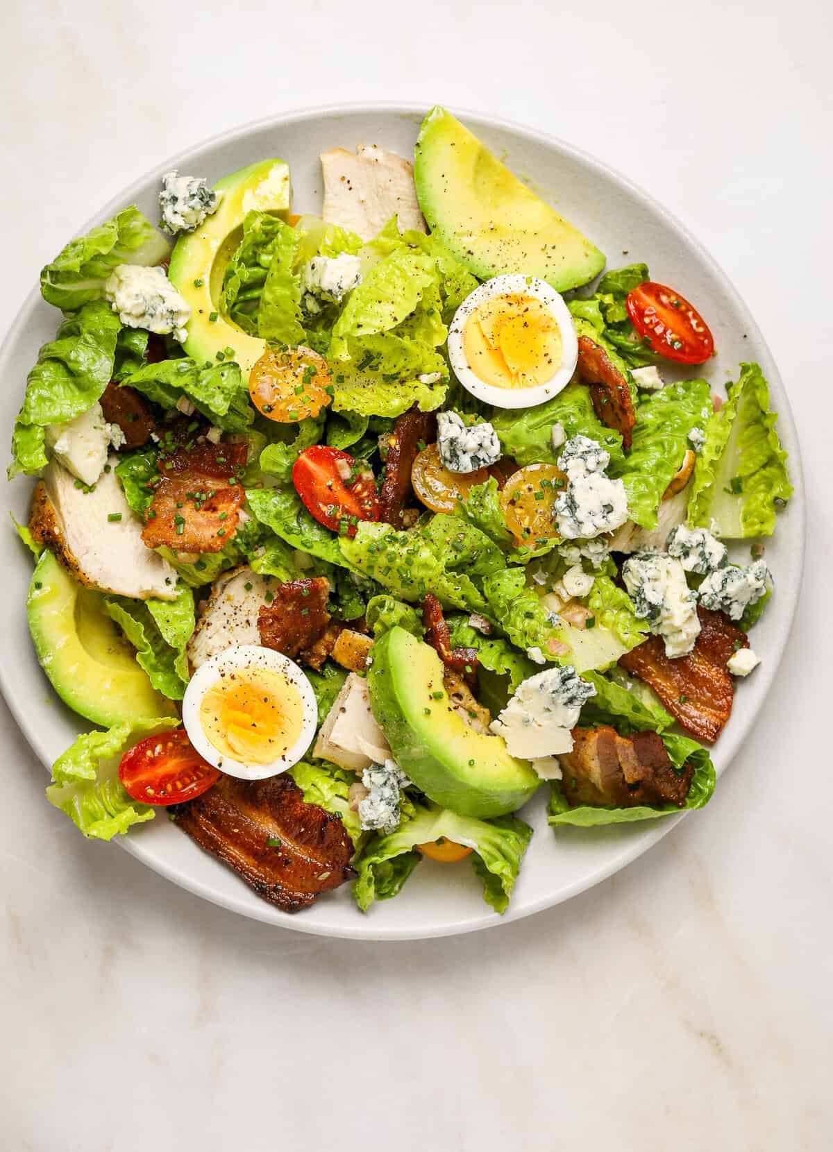 A white plate filled with salad, sliced tomatoes, avocado slices, chives and crumbled blue cheese