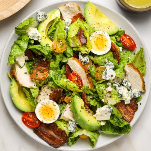 A plate of salad made up of hard boiled egg, lettuces leaves, bacon and halved tomatoes, avocado and cheese.