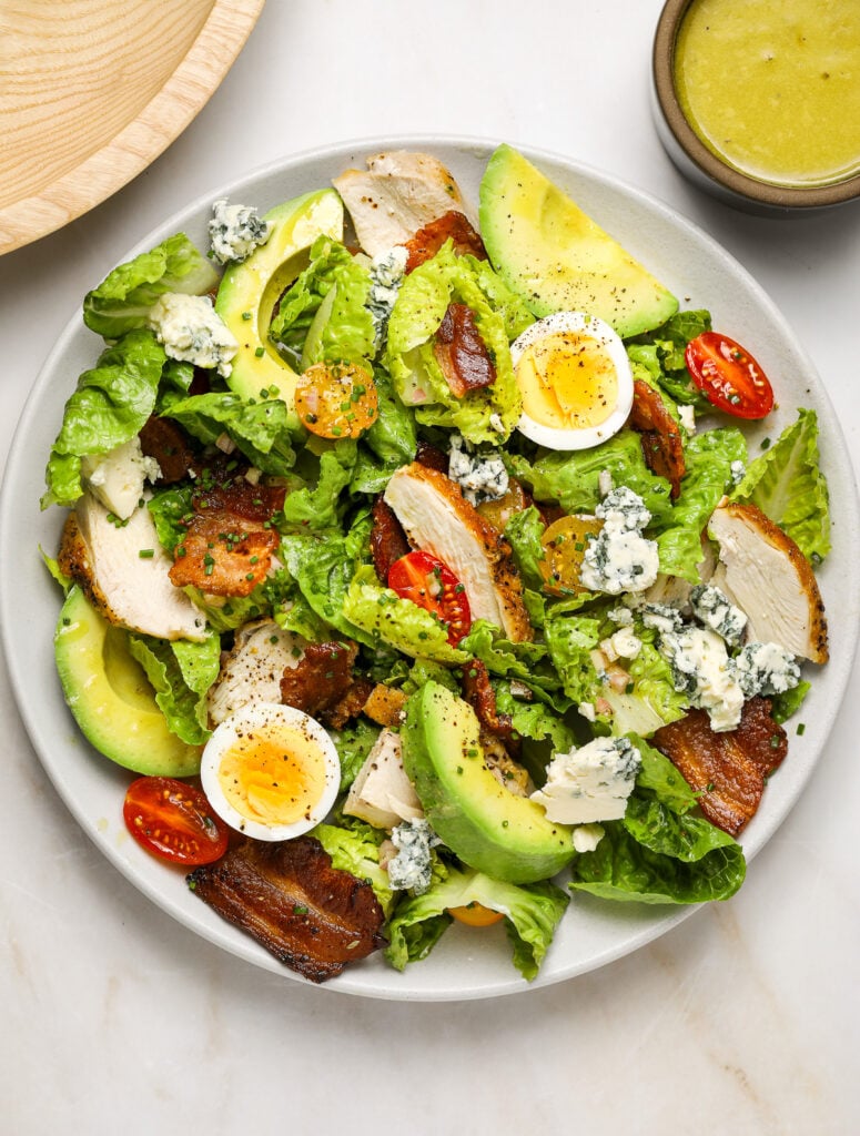 A plate of salad made up of hard boiled egg, lettuces leaves, bacon and halved tomatoes, avocado and cheese.