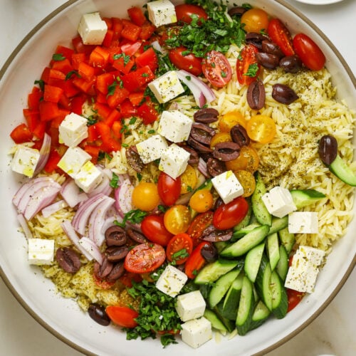 Orzo salad ingredients piled into a large ceramic bowl surrounded by a halved lemon, dried oregano, olive oil, a pepper mill and a dish of olives.