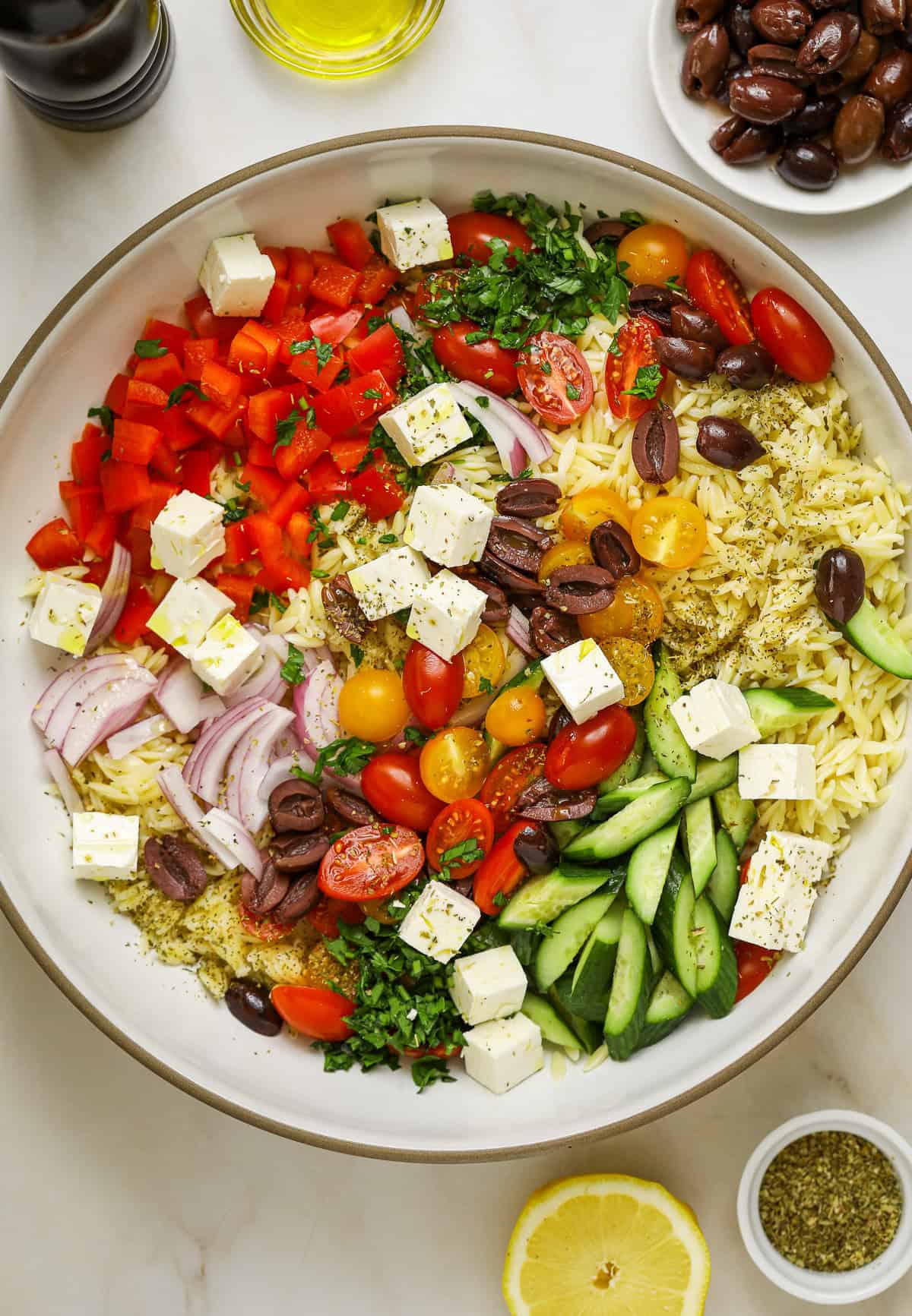 Orzo salad ingredients piled into a large ceramic bowl surrounded by a halved lemon, dried oregano, olive oil, a pepper mill and a dish of olives.