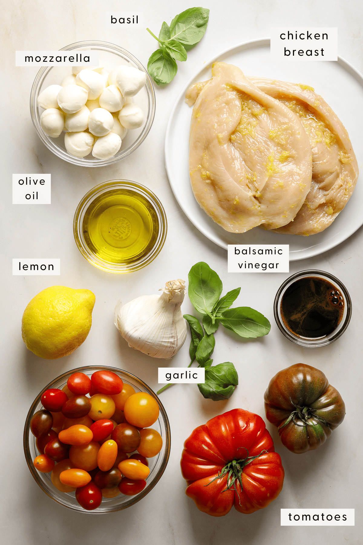 Recipe ingredients individually portioned: heirloom tomatoes, chicken breast, mozzarella balls, fresh basil, olive oil.