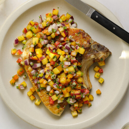 A large pork chip topped with peach salsa on a beige plate.