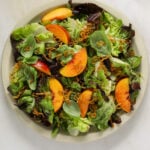 A large, round platter filled with salad, sliced peaches, fried shallots and whole basil leaves.