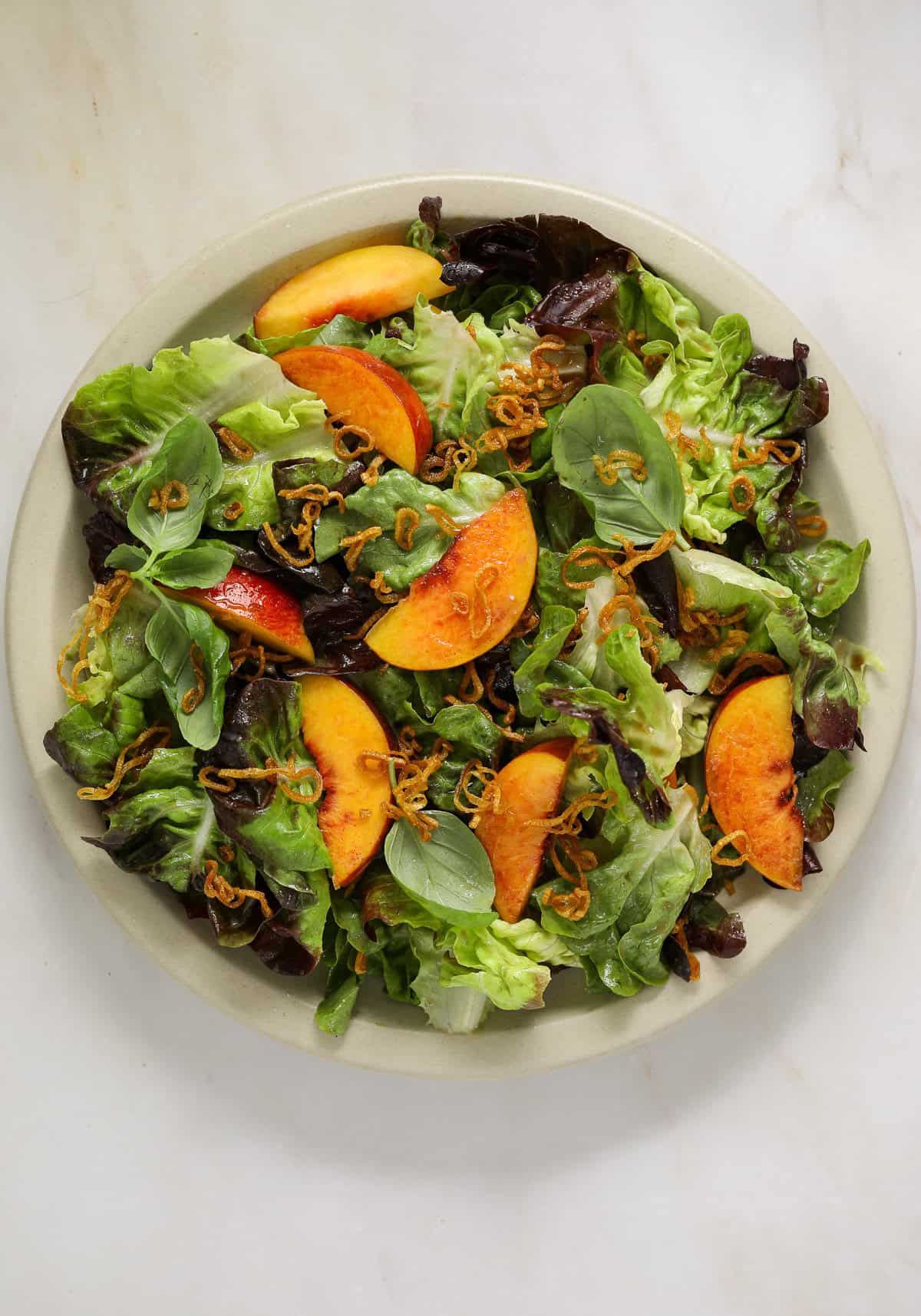 A large, round platter filled with salad, sliced peaches, fried shallots and whole basil leaves.