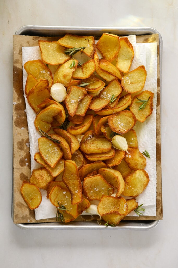 a paper towel lined baking tray filled with fried potatoes, garlic cloves and rosemary.