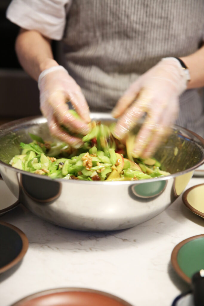 Gloved hands tossing a salad in a silver mixing bowl.