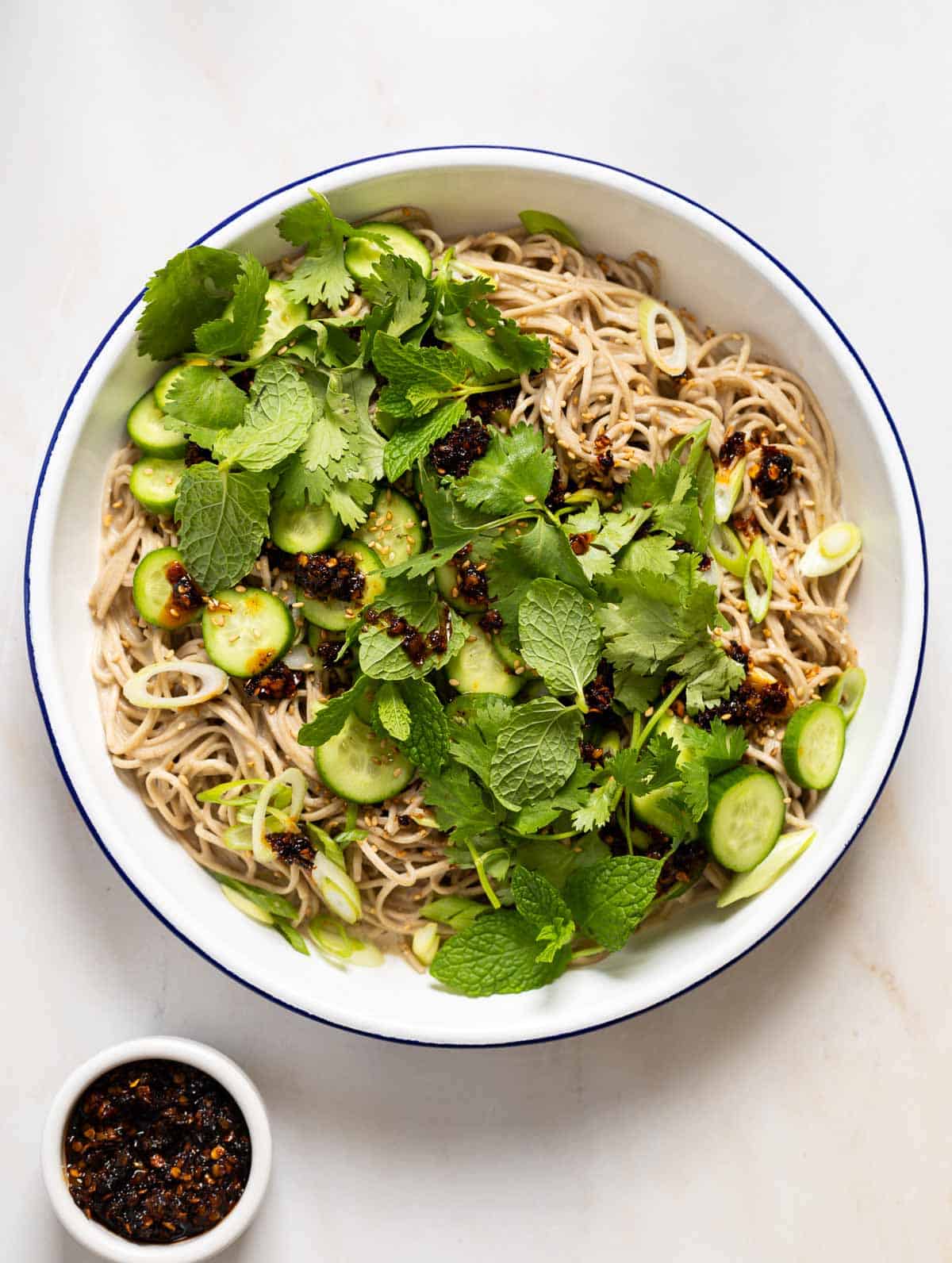 A white bowl with a blue rim filled with Asian noodles, cucumber, mint and cilantro.