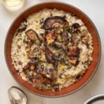 A brown, ceramic bowl of risotto topped with roasted mushrooms. Two wine glasses at the top of frame and spoons off to the left.
