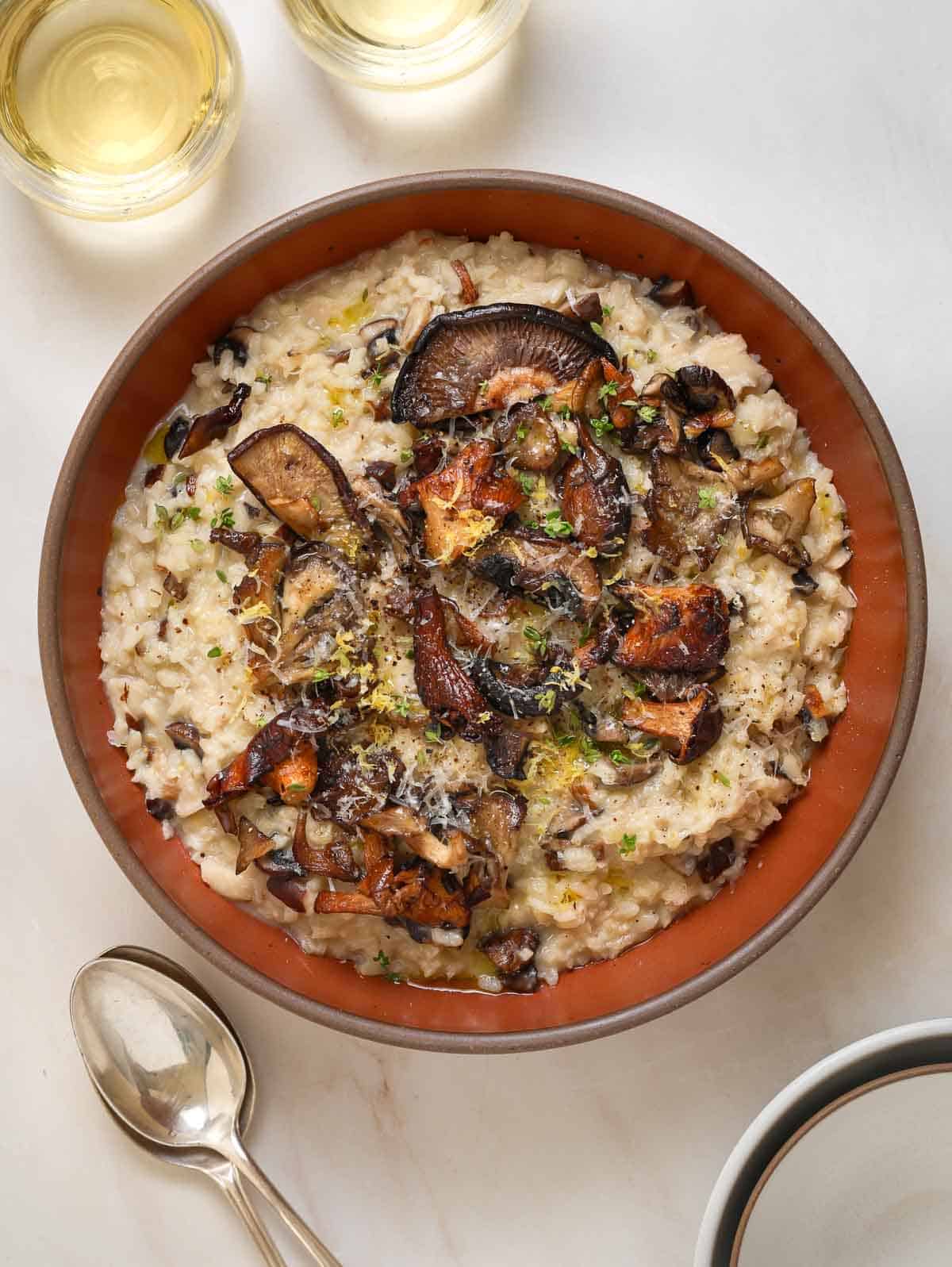 A brown, ceramic bowl of risotto topped with roasted mushrooms. Two wine glasses at the top of frame and spoons off to the left.