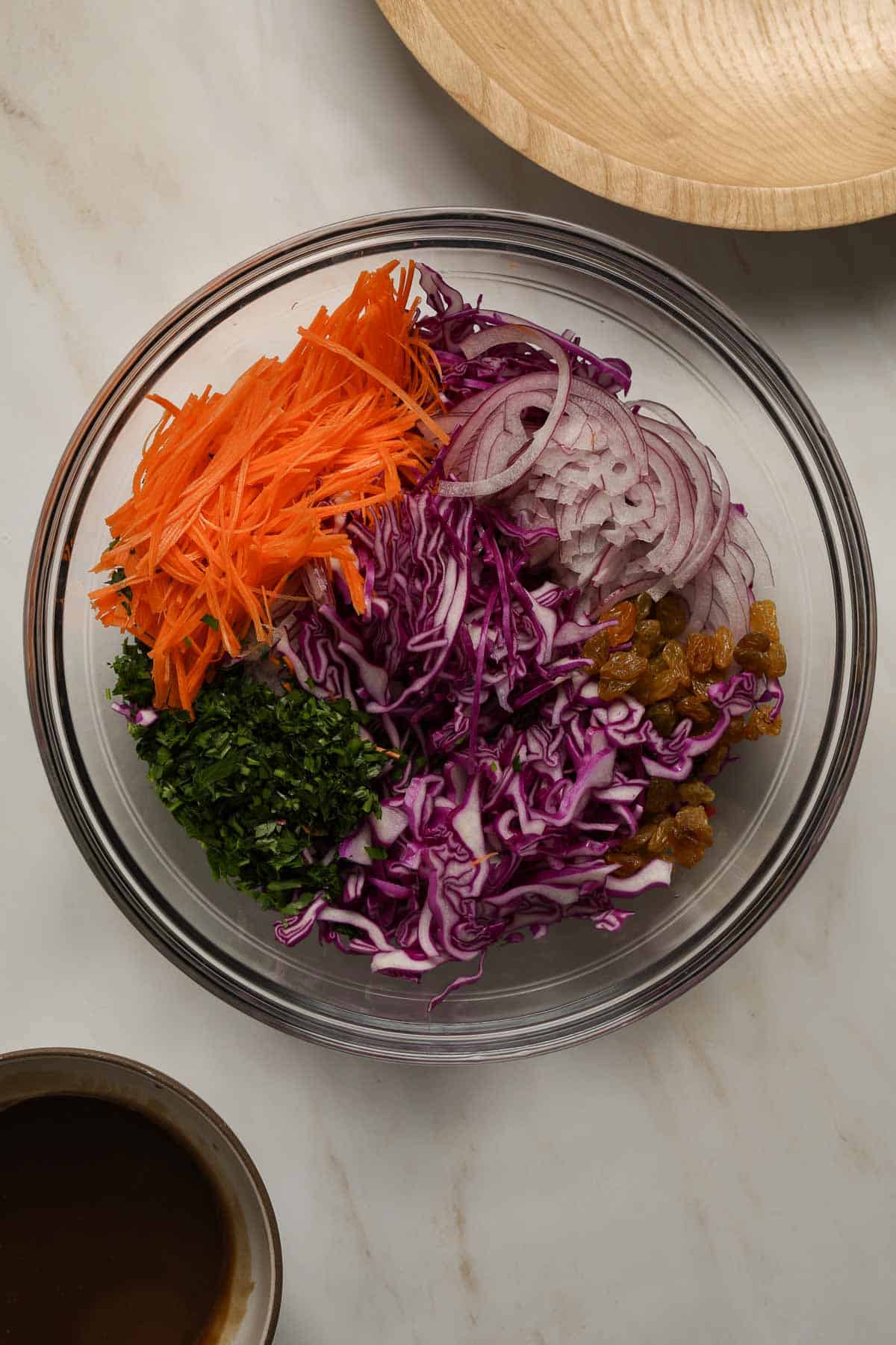 A glass mixing bowl filled with thinly shaved cabbage, carrot, onion and chopped parsley.