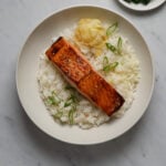A white ceramic bowl filled with sushi rice, a filet of salmon and pickled ginger.