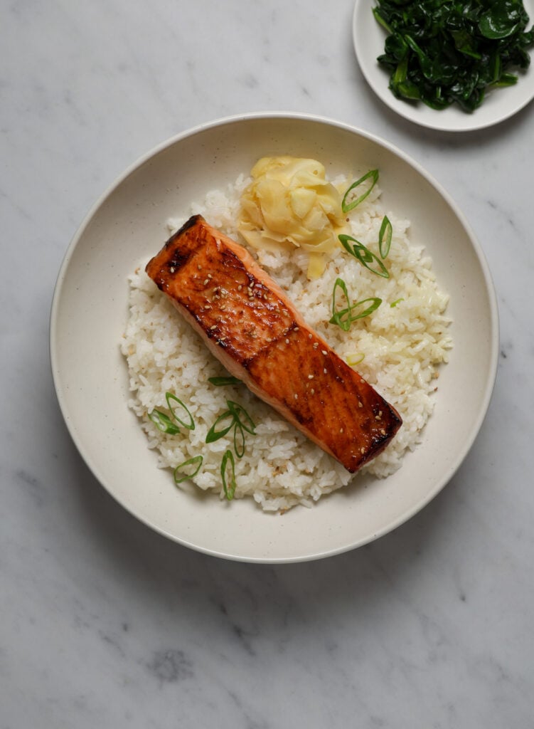 A white ceramic bowl filled with sushi rice, a filet of salmon and pickled ginger.