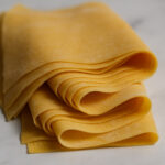 Thin, yellow sheets of fresh pasta dough gently folded on a marble tabletop.