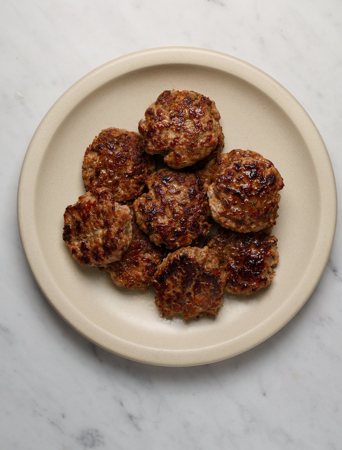 A round, ceramic platter filled with a pile of glistening sausage patties on a marble tabletop.