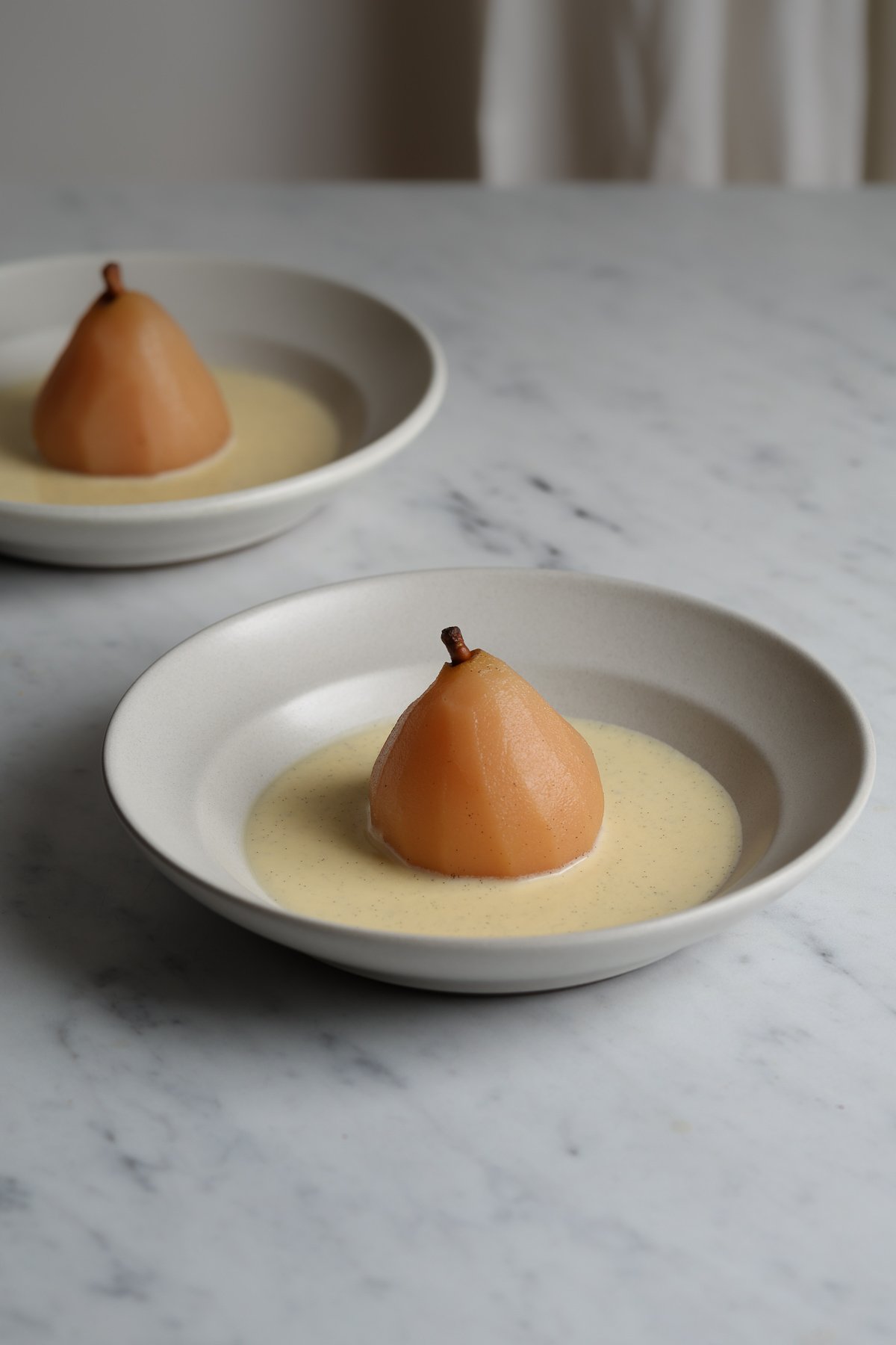 Two poached pears, in ceramic bowls, surrounded by vanilla sauce.