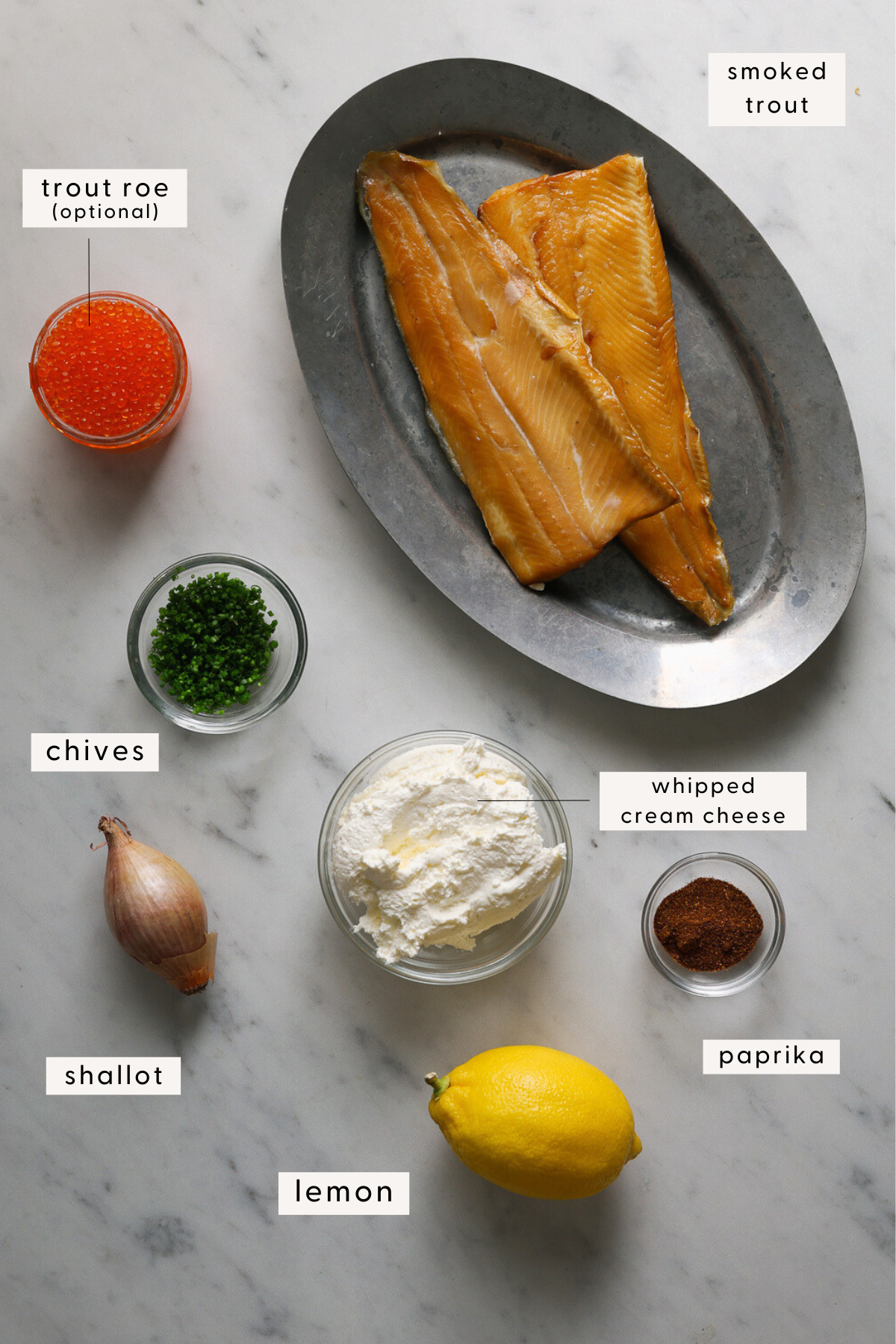 Smoked trout on a silver platter, minced chives, a bowl of cream cheese, a lemon, and paprika on a marble tabletop.