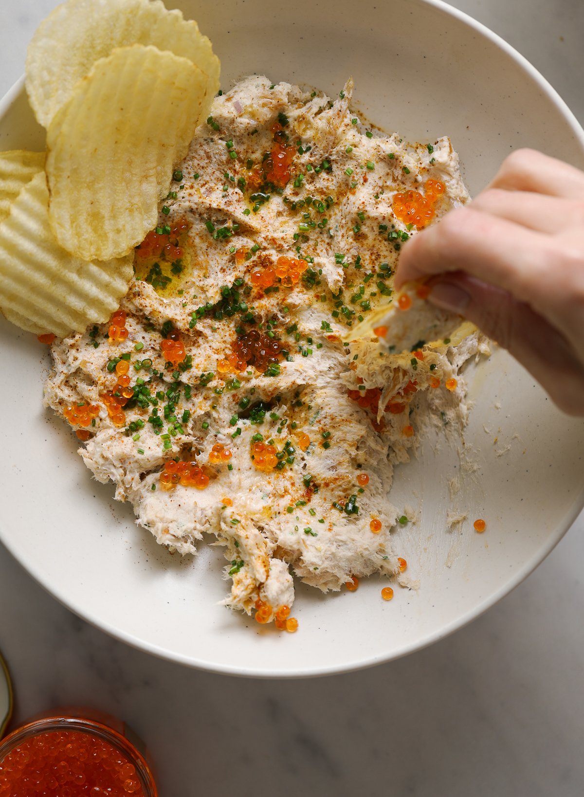 A hand scooping up a bite of dip on a ruffled potato chip.