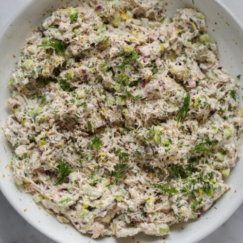 A bowl of tuna salad topped with dill and celery on a marble table top.