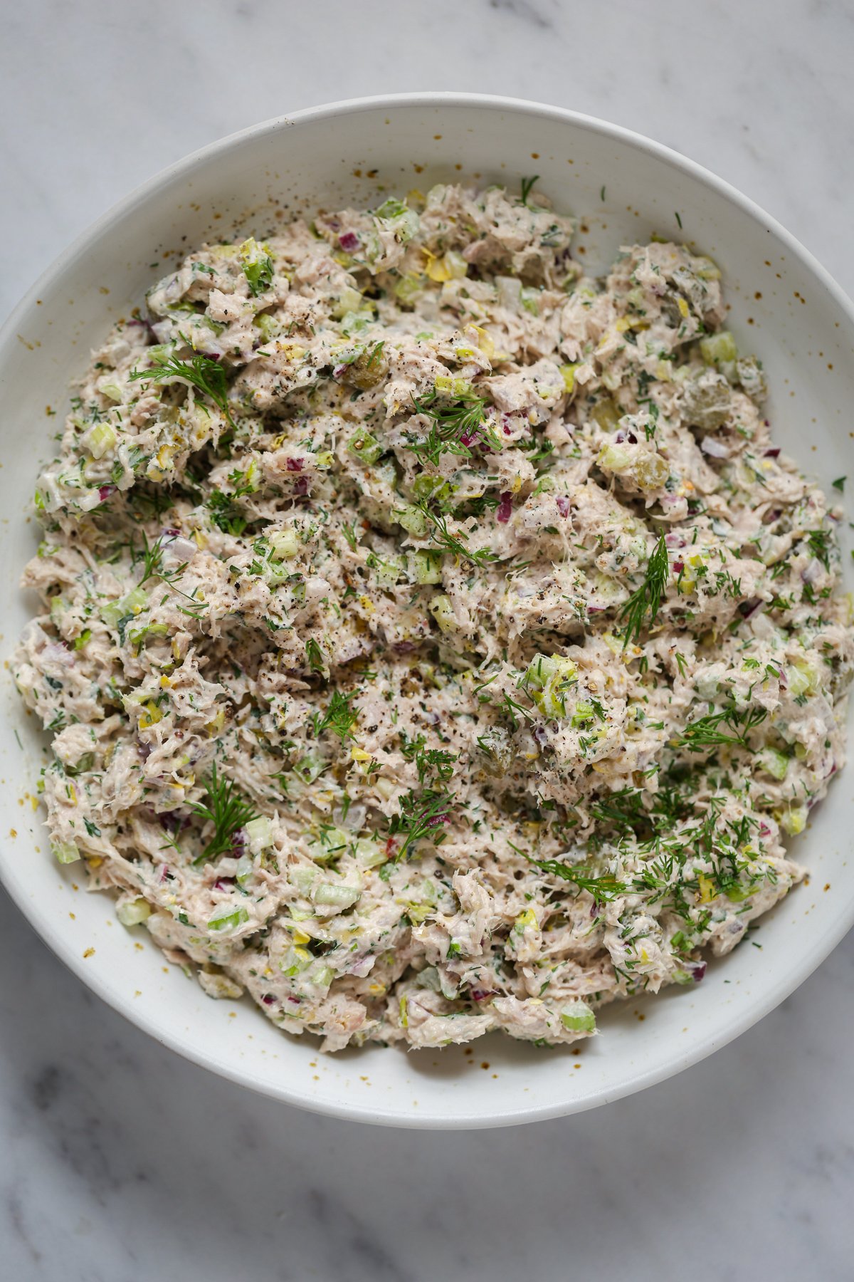 A bowl of tuna salad topped with dill and celery on a marble table top.