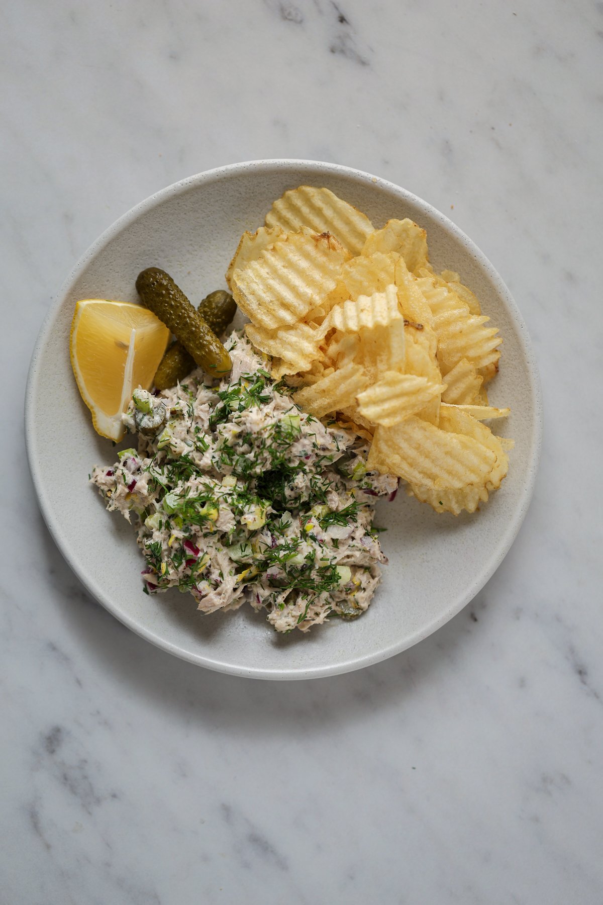 A pile of salad on a plate with potato chips, cornichons and lemon.